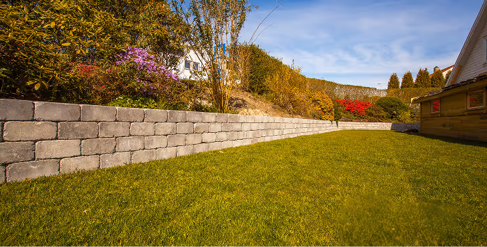 Garten mit grünem Rasen, einer Stützmauer aus grauen Steinen und bunten Sträuchern unter blauem Himmel.