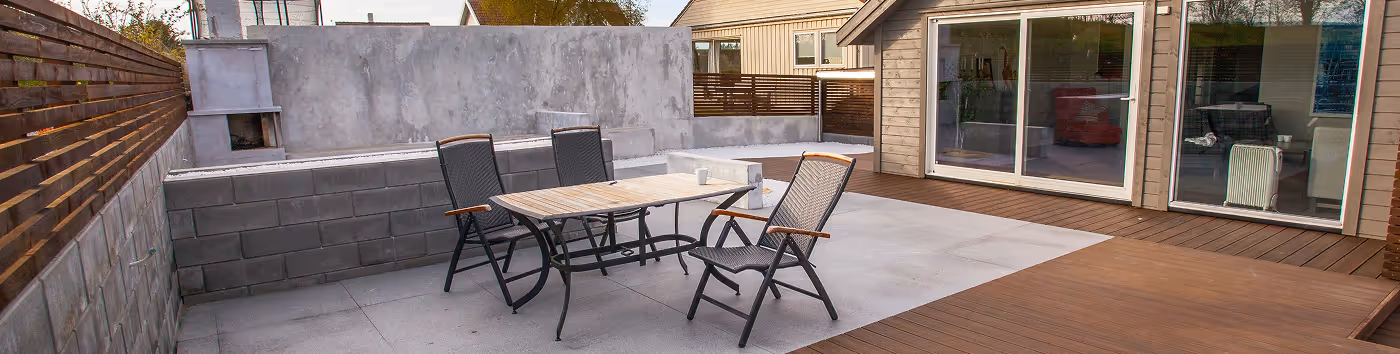 Moderne Terrasse mit grauen Fliesen, Holzboden, einem Tisch mit drei Stühlen und Glas-Schiebetüren an einem Haus.