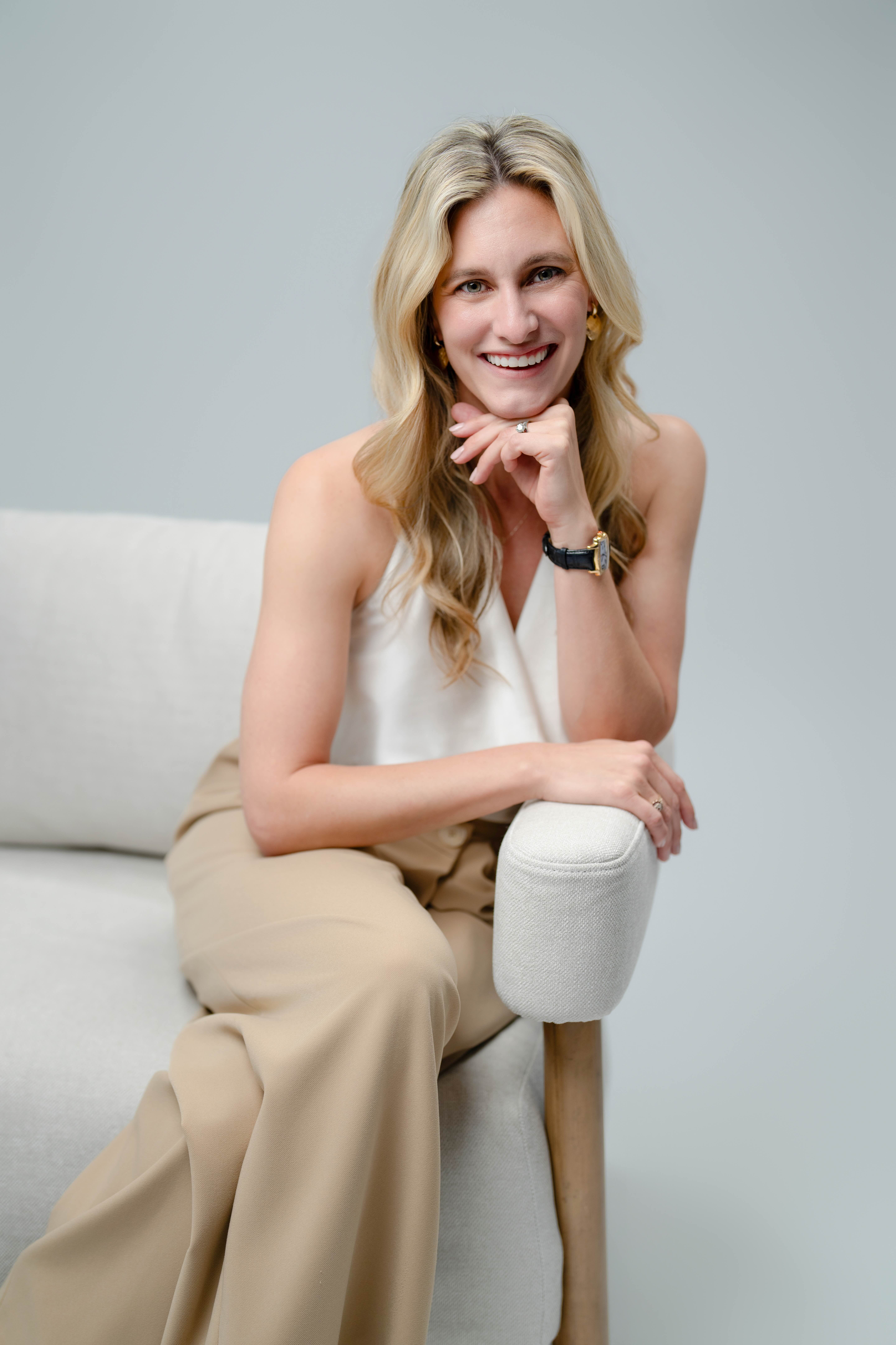 Smiling blonde woman sitting on a light gray armchair with a relaxed pose against a plain gray background.
