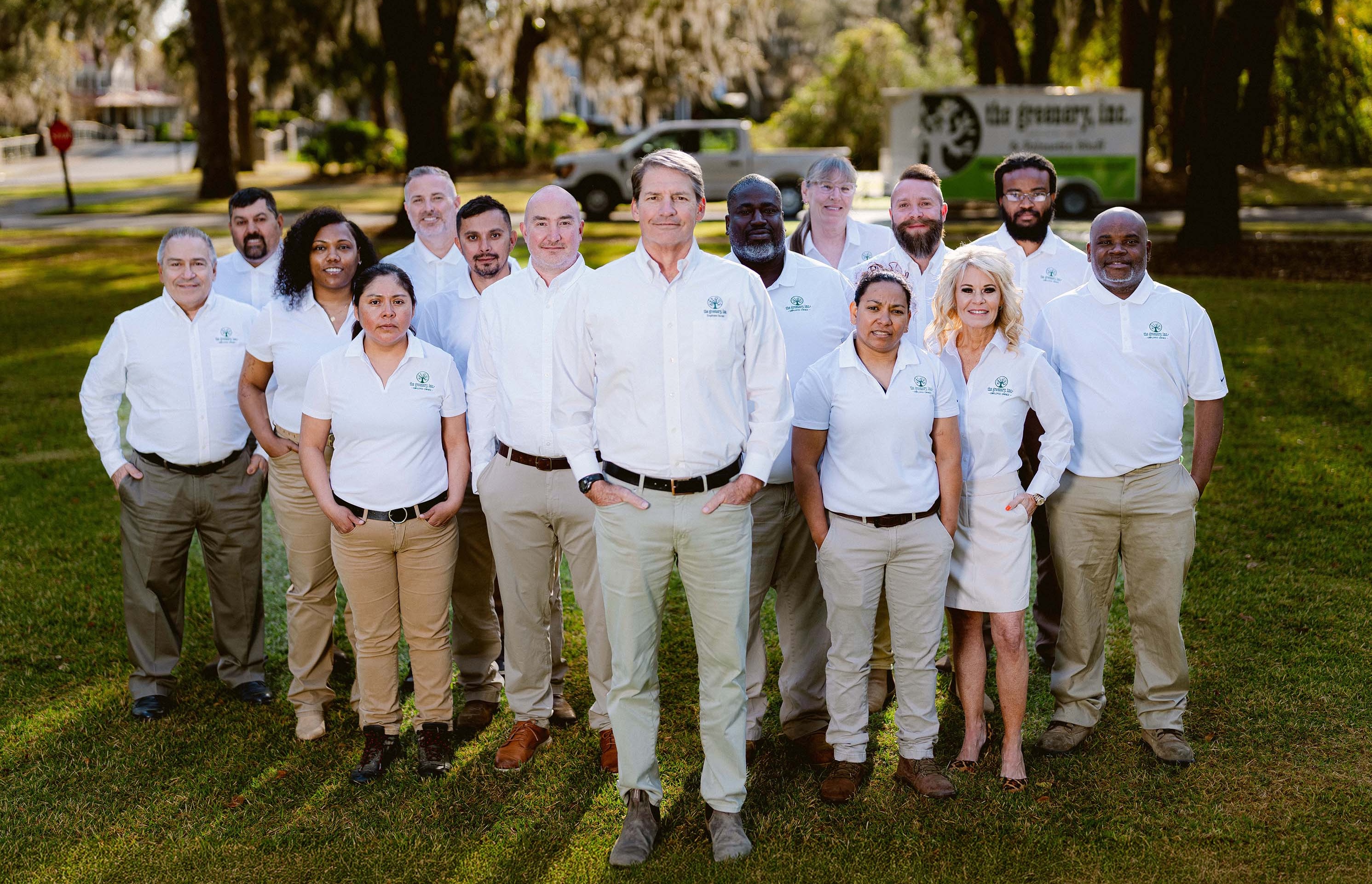 Group of 15 diverse employees outdoors wearing white company shirts and khaki pants, standing on grass with trees in background.