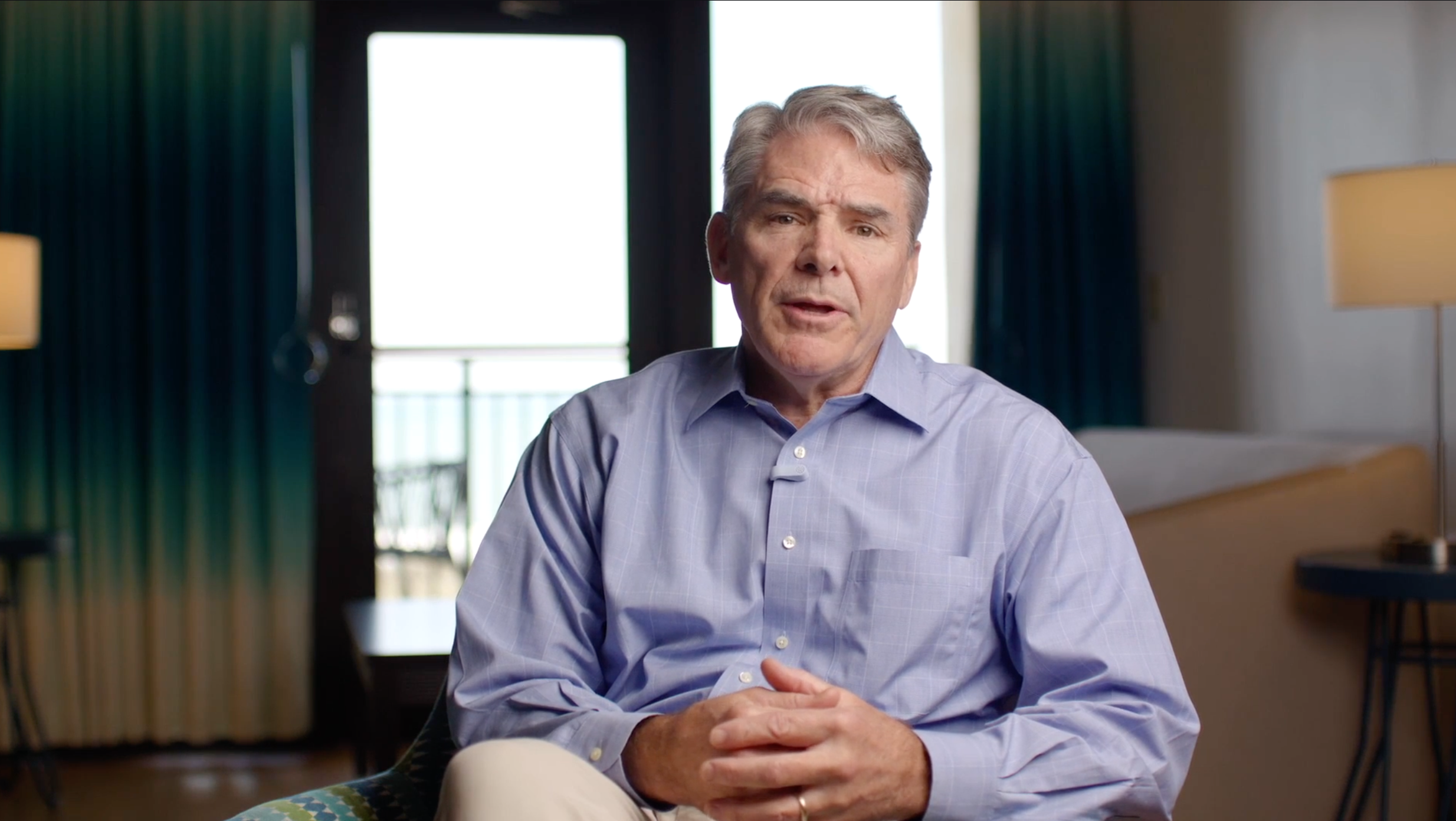 Middle-aged man in a blue button-up shirt sitting and speaking in a softly lit room with curtains and lamps in the background.