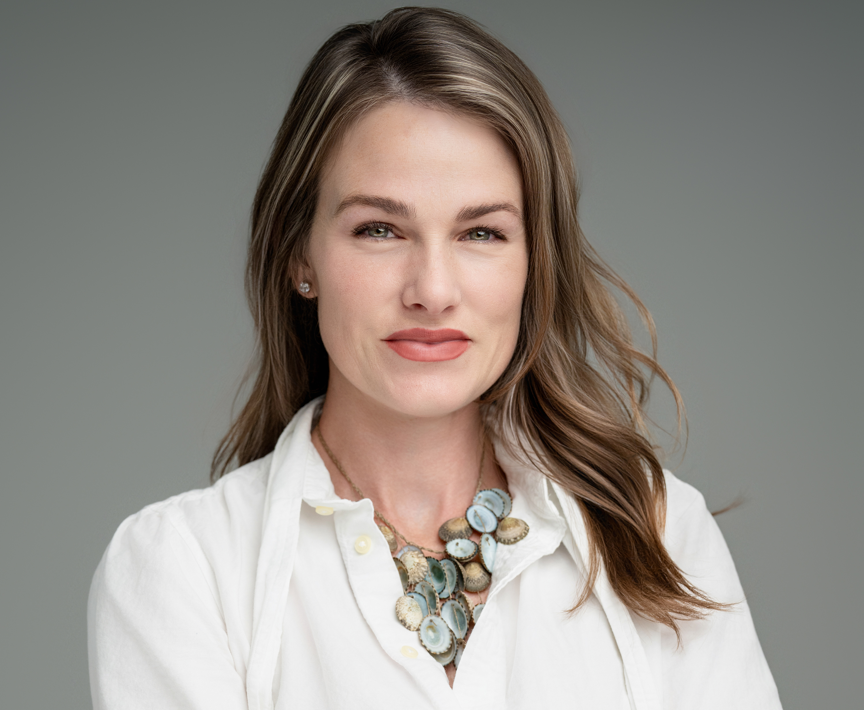Portrait of a woman with light brown hair wearing a white shirt and a multi-disc necklace against a gray background.
