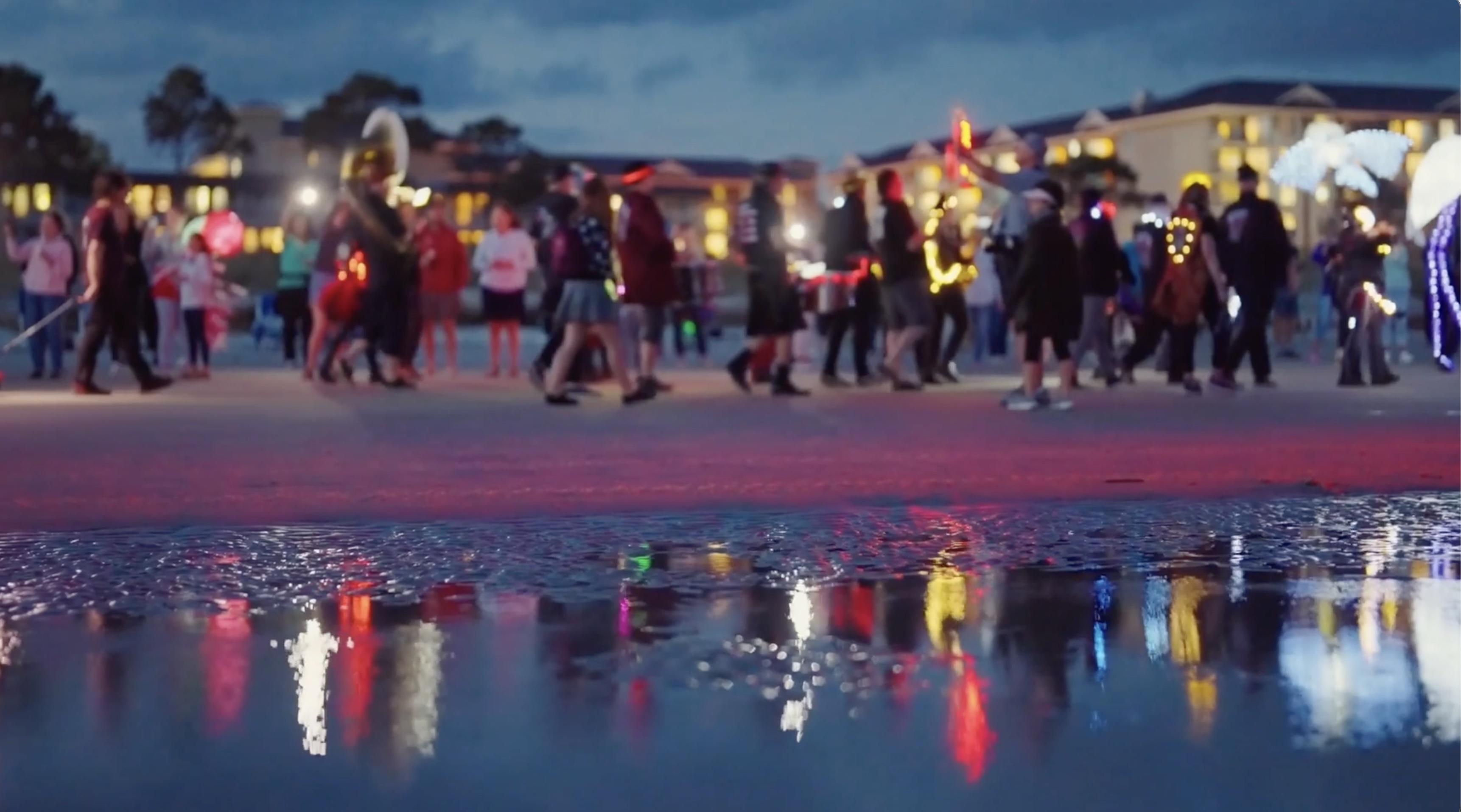 People walking in a nighttime parade with illuminated decorations reflected in a nearby puddle.