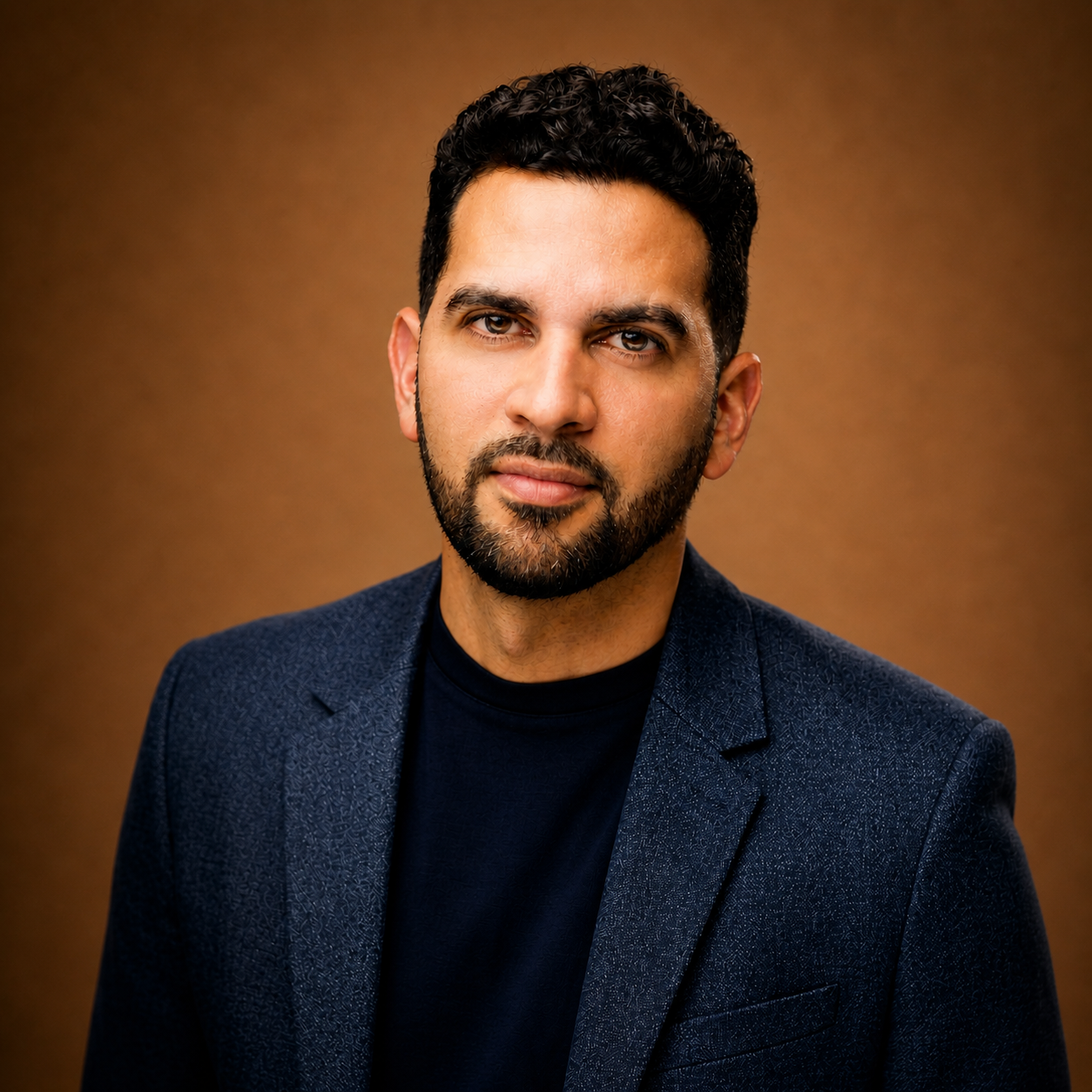 Smiling man with a beard wearing a black hat and black jacket against a dark background.
