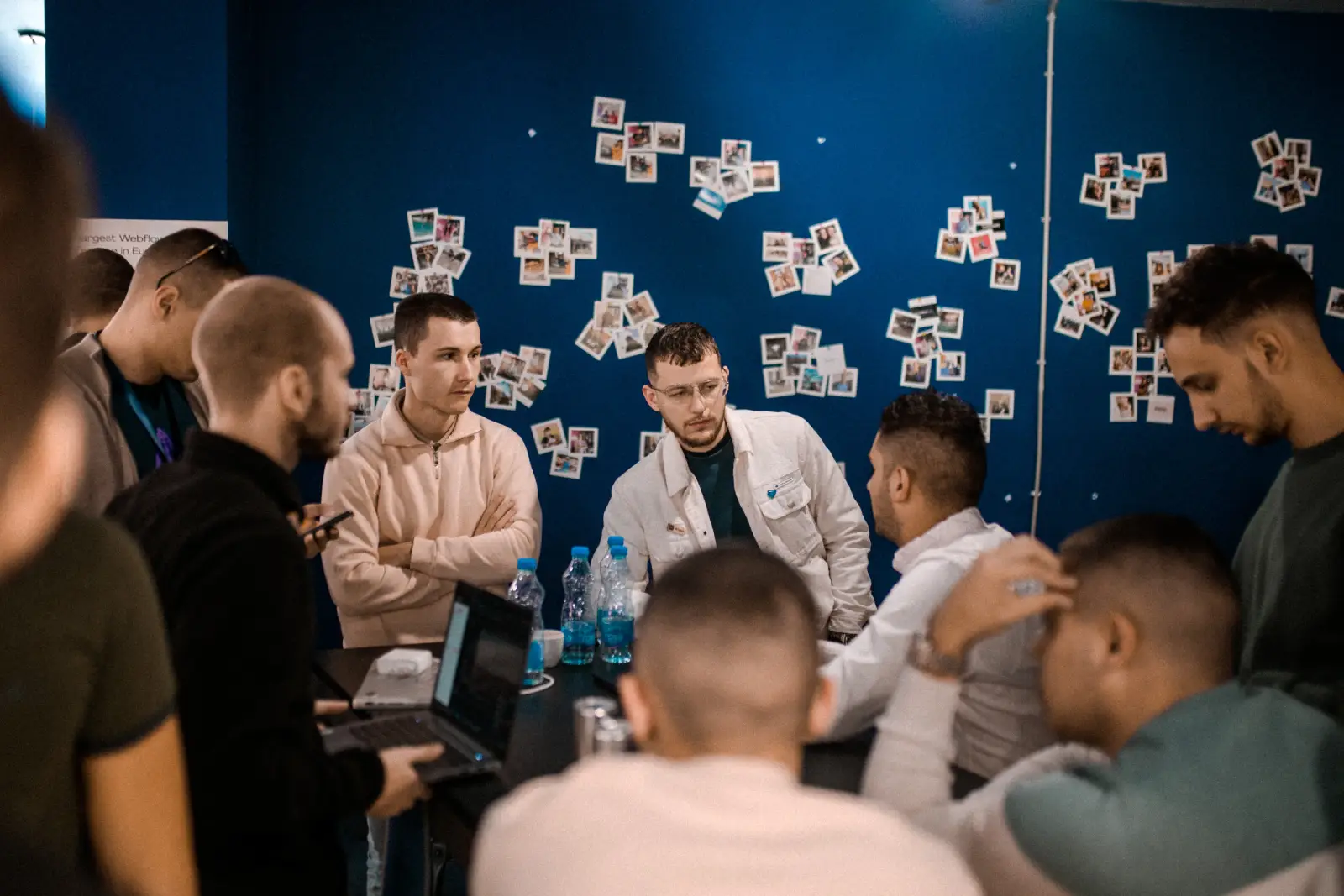 Group of men gathered around a table with laptops and water bottles in a room with blue walls and photos.
