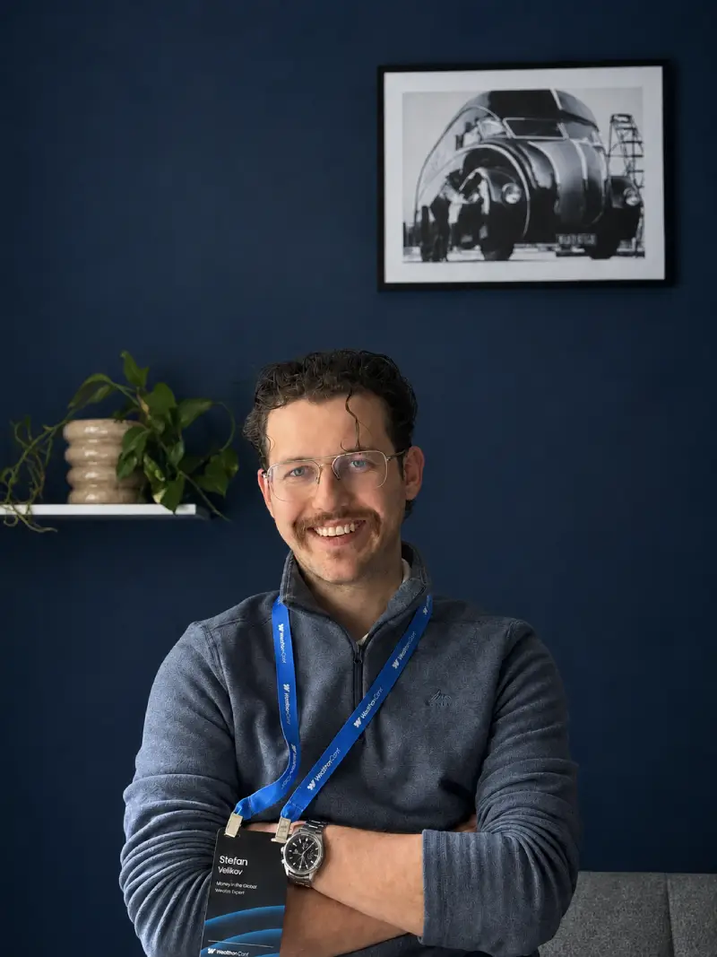 Smiling man with glasses and a mustache wearing a gray sweater and conference lanyard crossed arms.