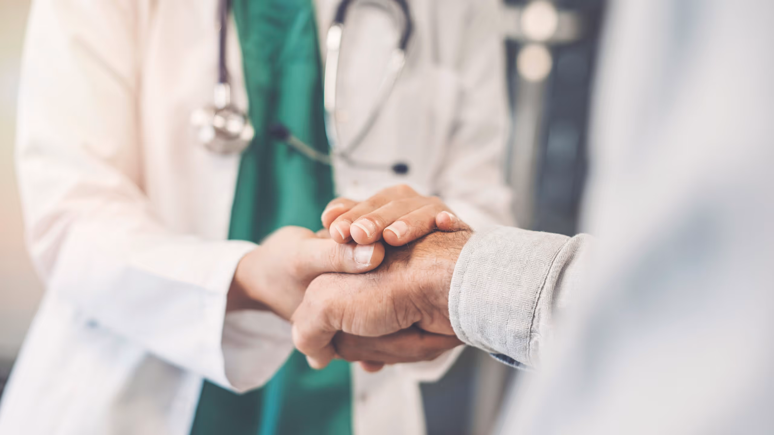 Doctor in white coat and green scrubs gently holding and comforting a patient's hand.