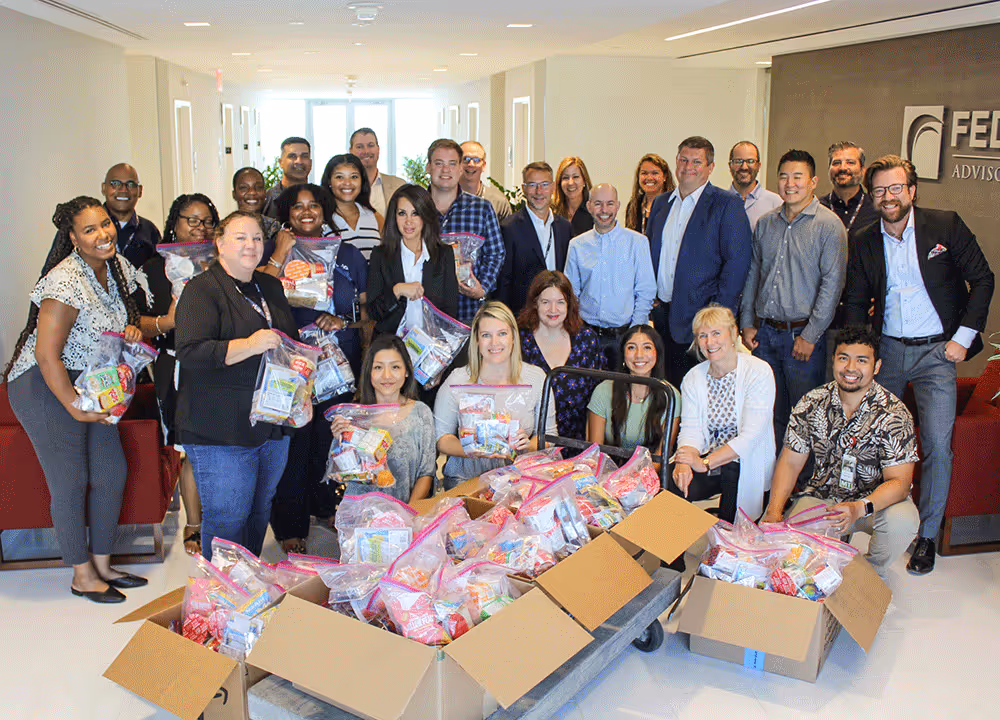 Group of diverse employees smiling and holding bags of donated food items gathered around boxes filled with more food packages in an office lobby.