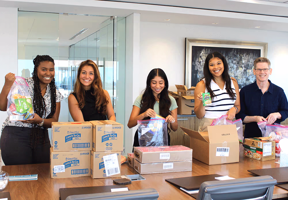 Five people smiling and packing food items into plastic bags and boxes in a bright office setting.