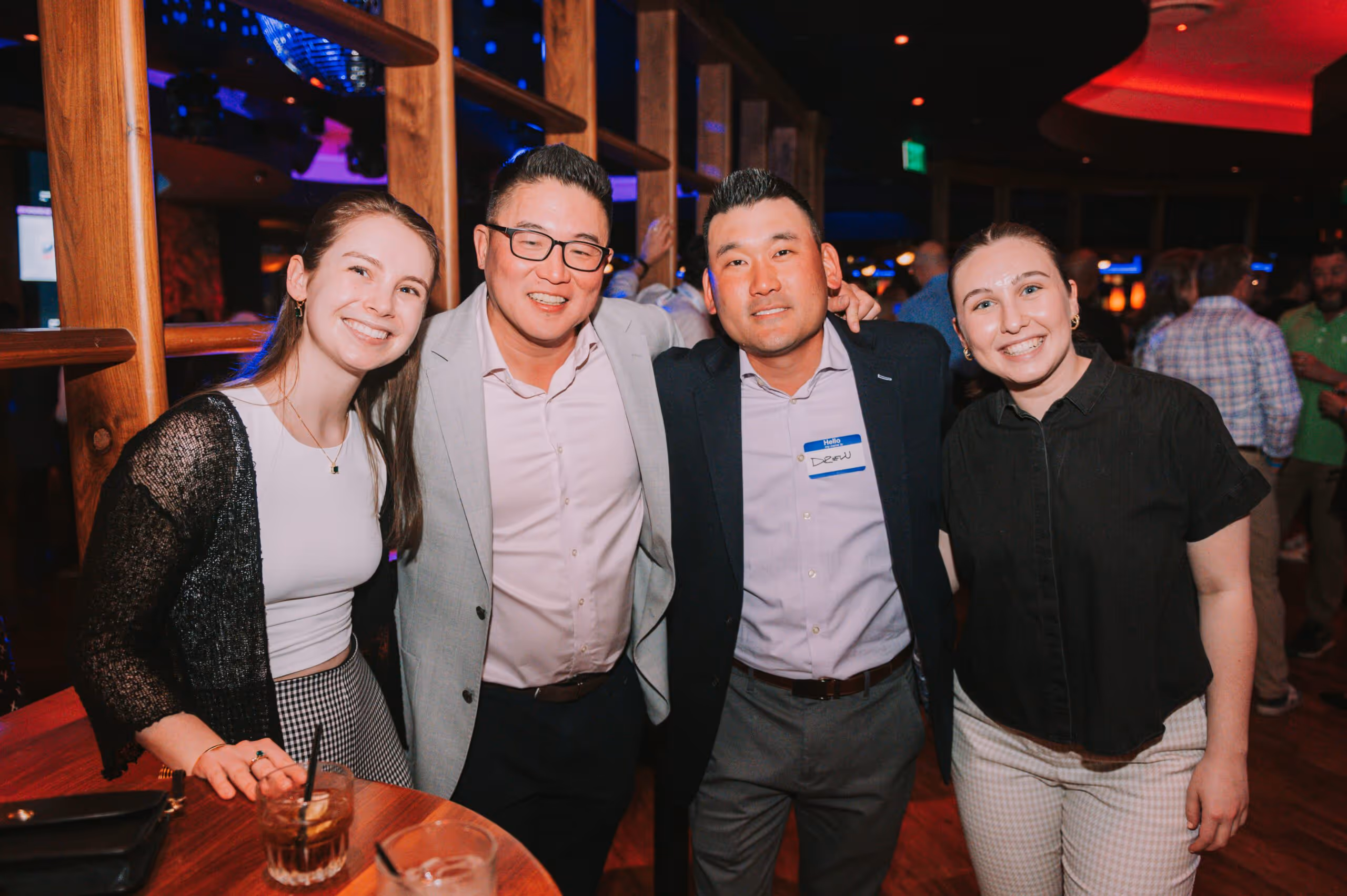Four smiling adults standing close together at an indoor social event with wooden decor and warm lighting.