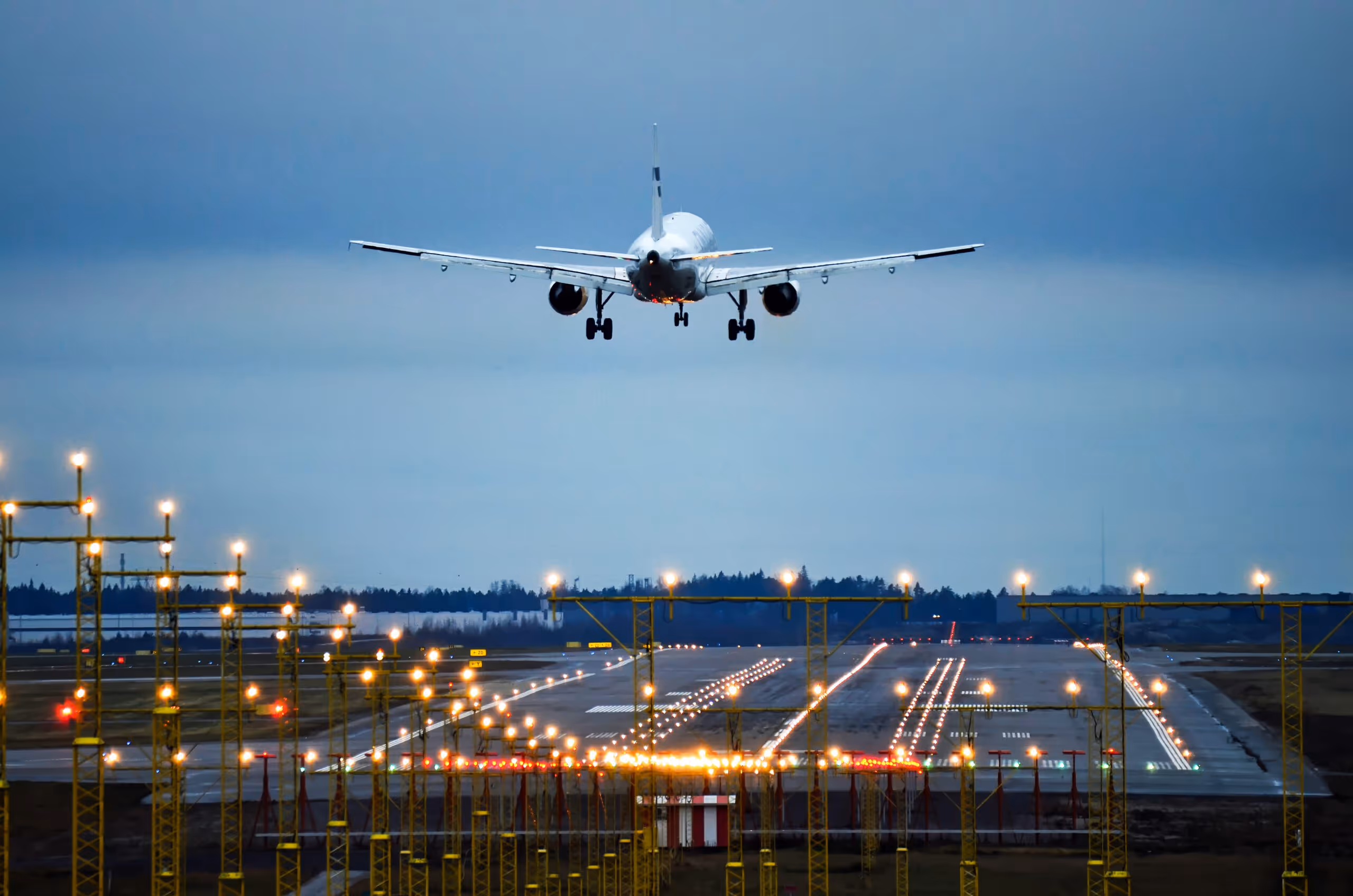 Airplane descending toward a lit runway at dusk with runway lights and guidance structures visible.
