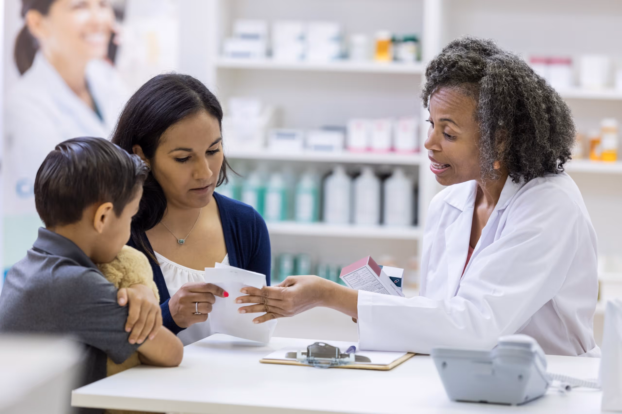 Pharmacist explaining medication instructions to a woman and a young boy holding a teddy bear in a pharmacy.