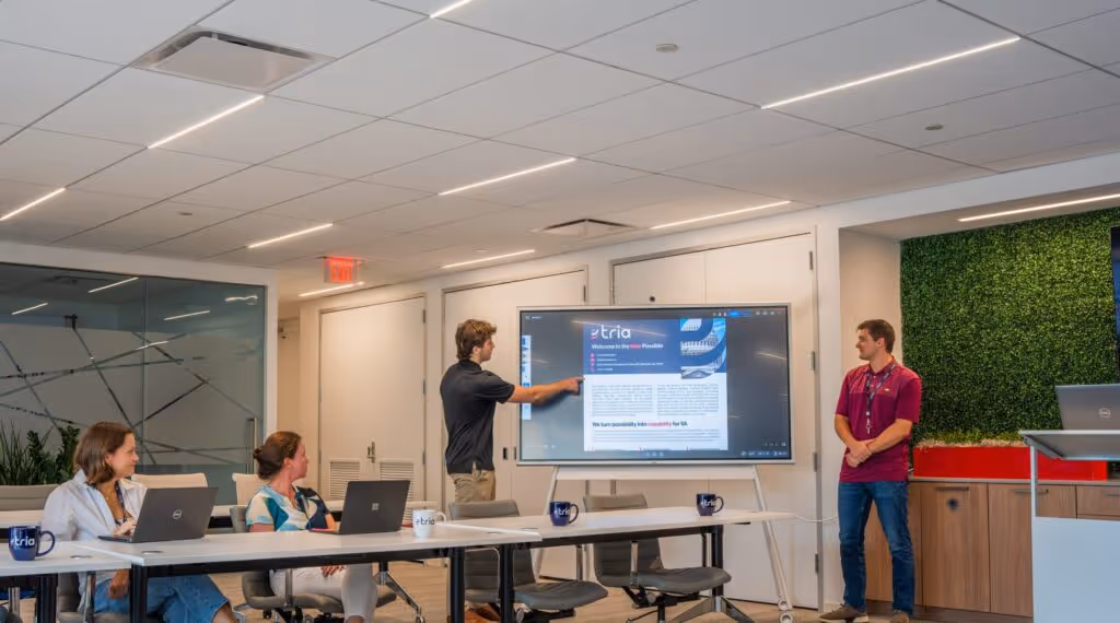 Two men standing and presenting information on a large screen while two women seated at a table watch attentively in a modern office conference room.