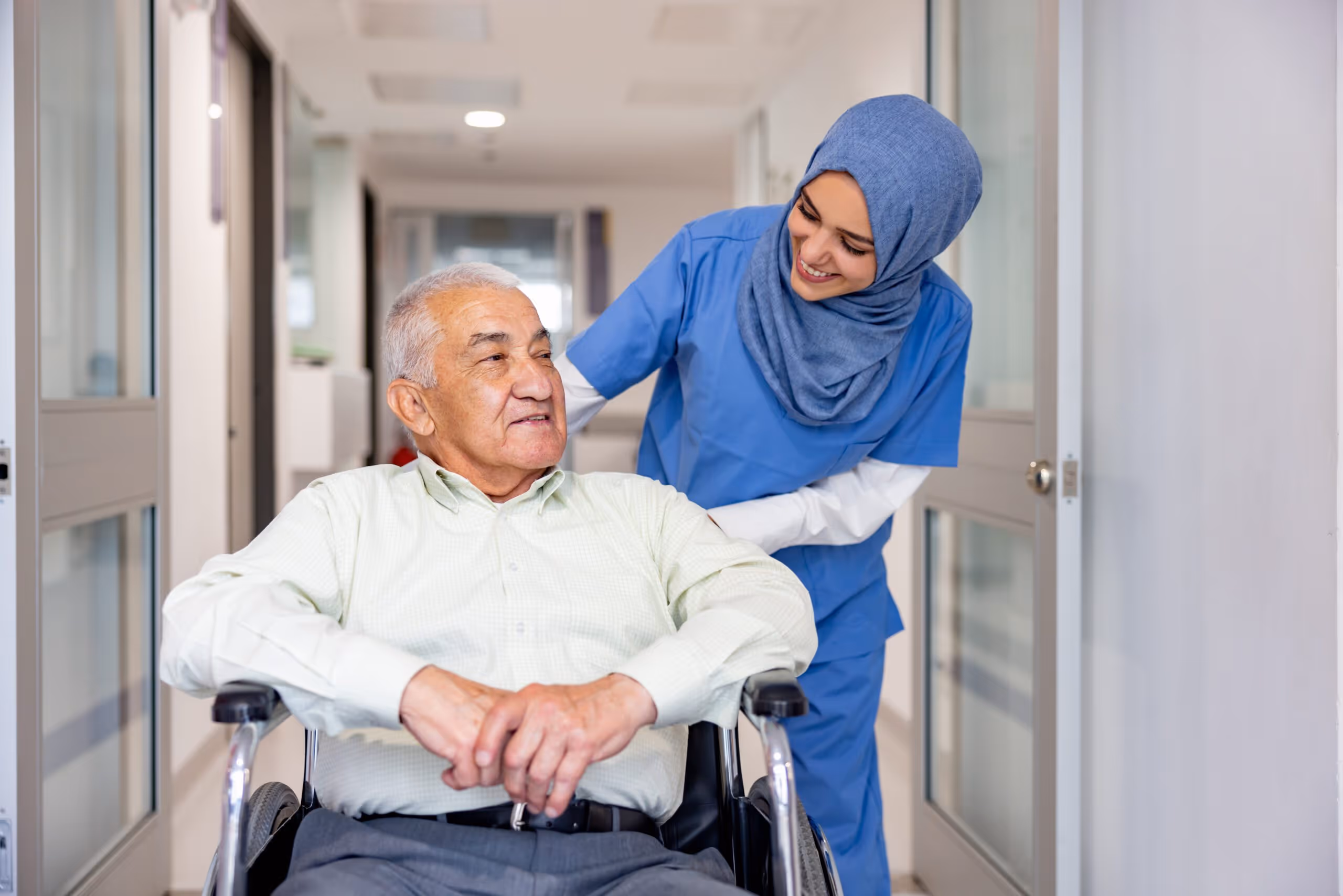 Smiling nurse in blue scrubs and hijab pushing an elderly man in a wheelchair down a hospital corridor.