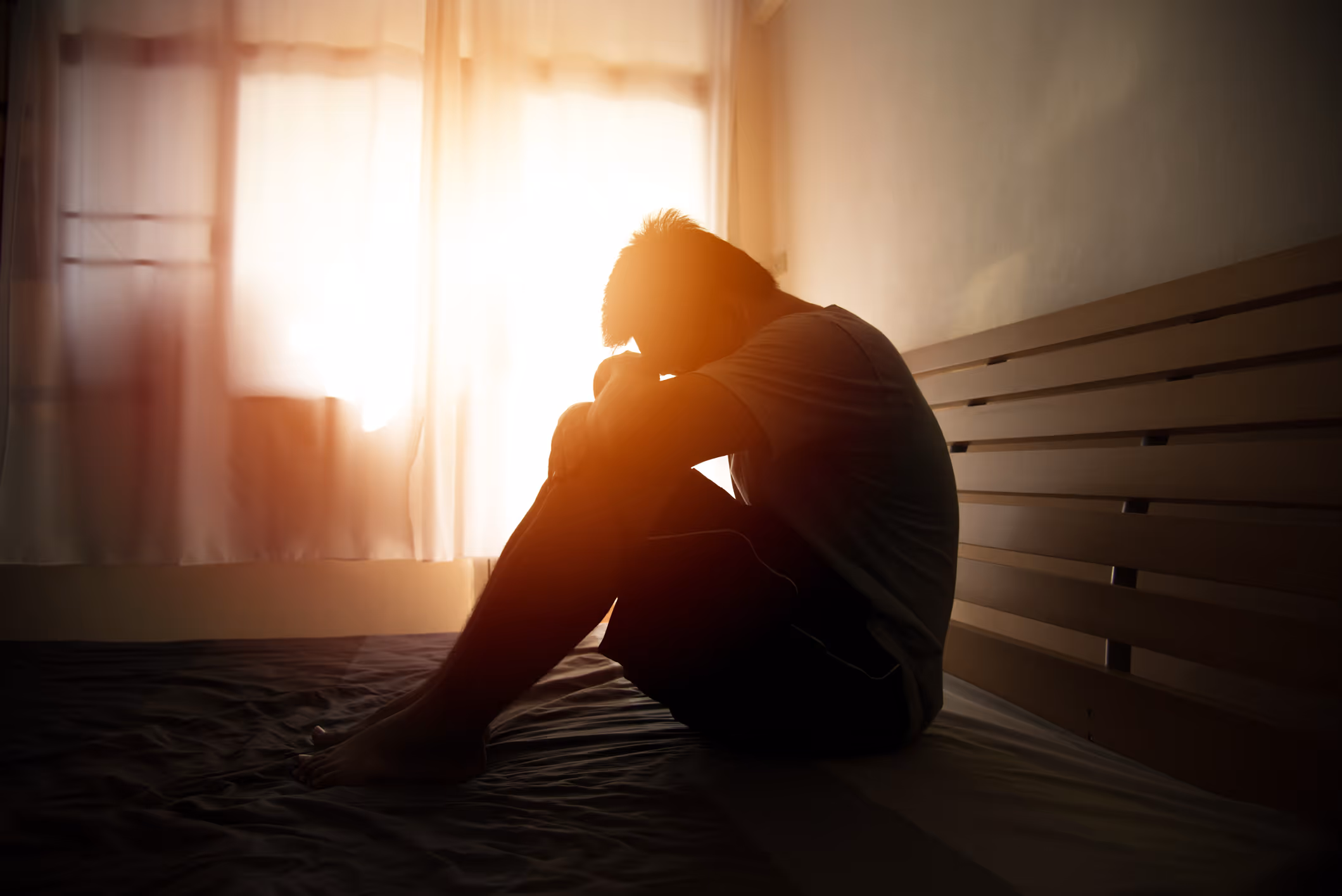 Silhouette of a person sitting on a bed with knees drawn up and head resting on arms, backlit by bright window light.