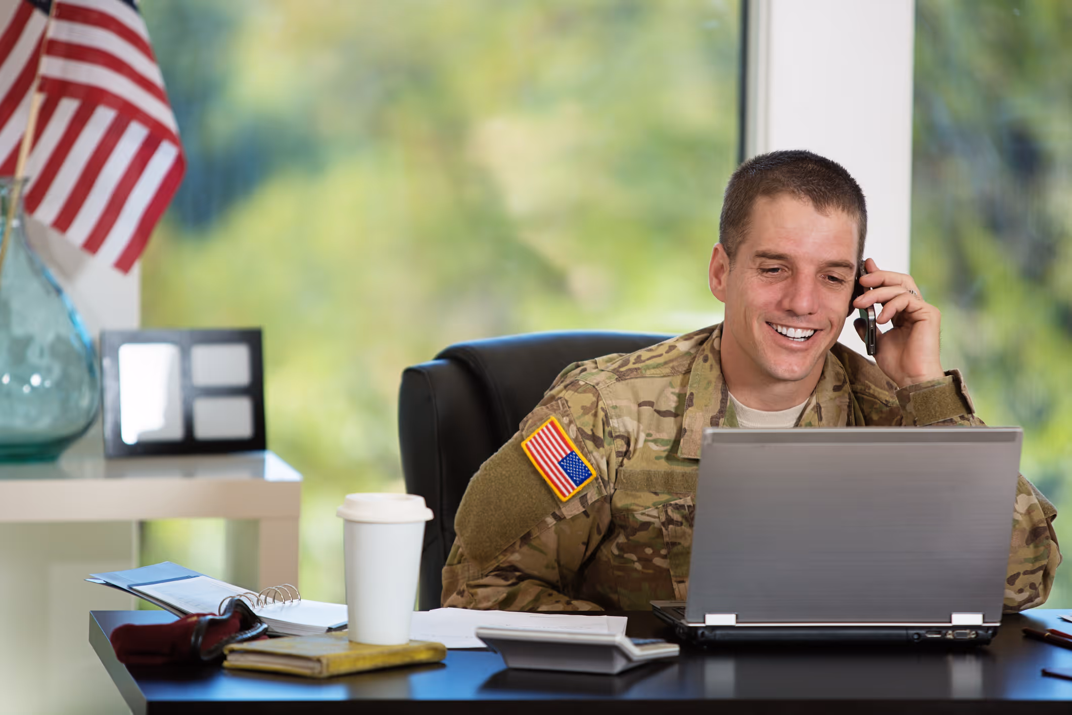 Smiling male soldier in camouflage uniform talking on a phone while working on a laptop at a desk with an American flag in the background.