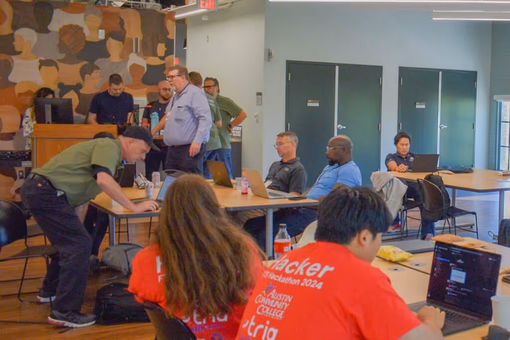 People working on laptops in a large room during a hackathon event, with some wearing red hacker t-shirts.