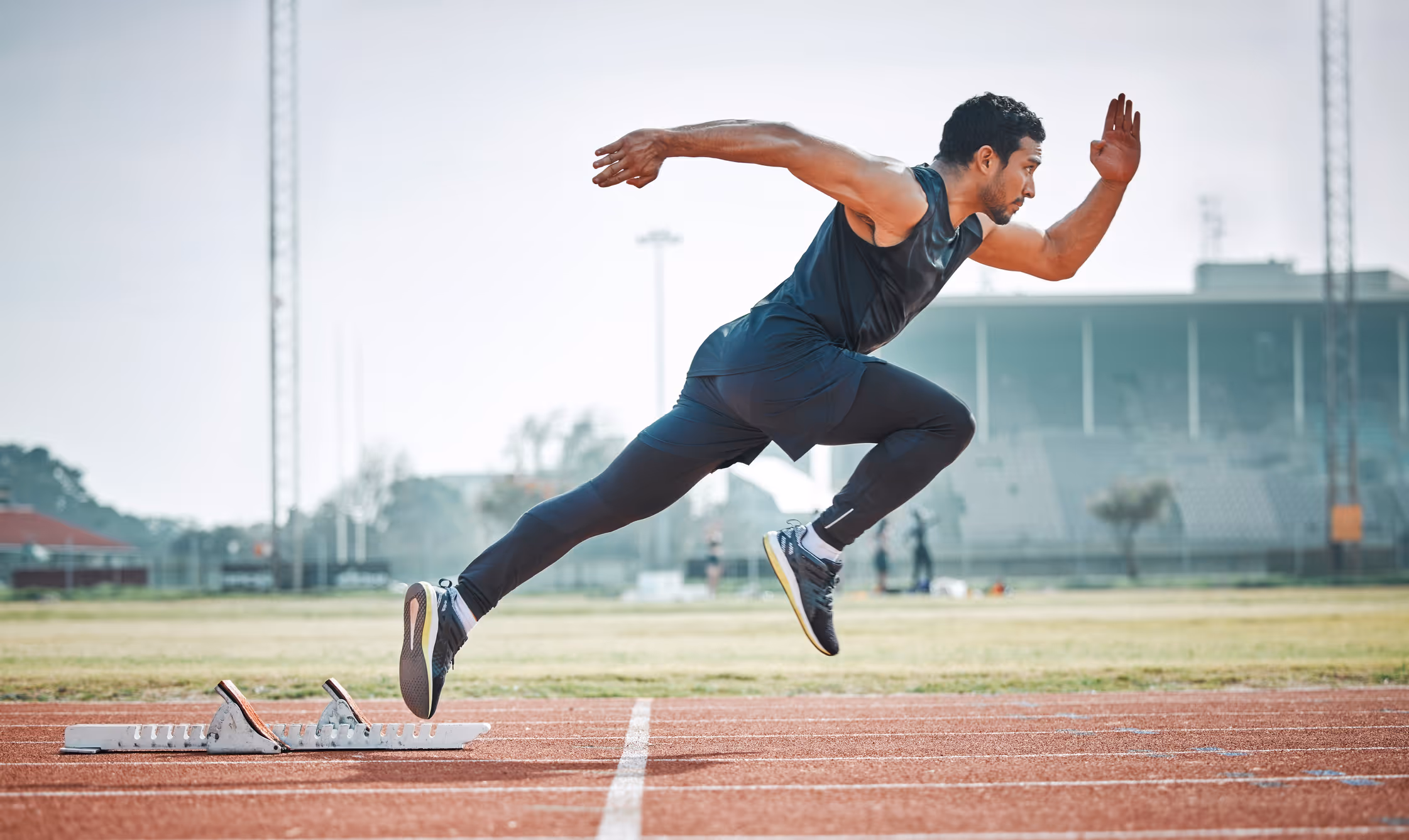Athlete in black sportswear sprinting off starting blocks on an outdoor track.
