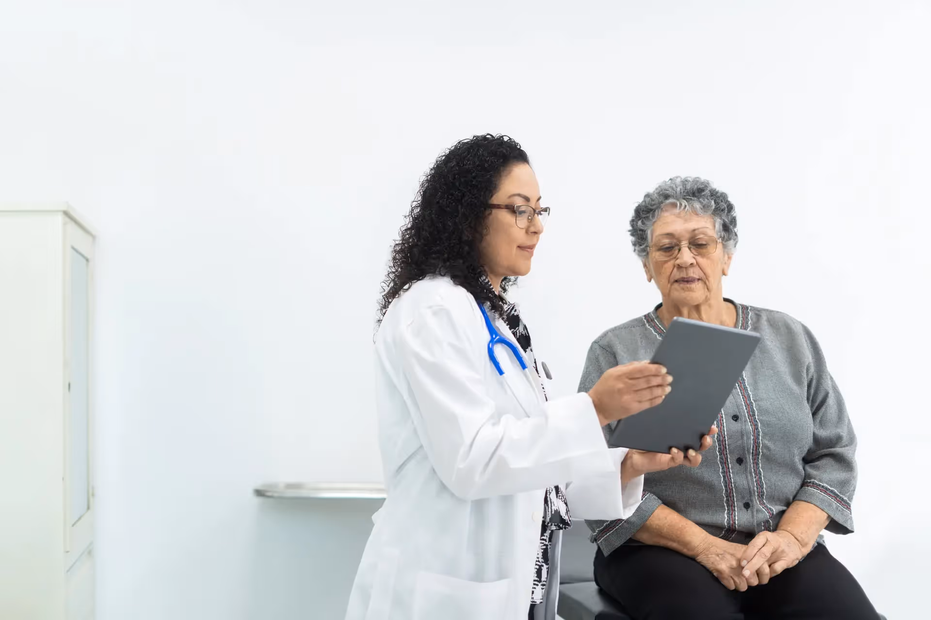 Female doctor showing a tablet to an elderly woman sitting on an exam table in a medical office.