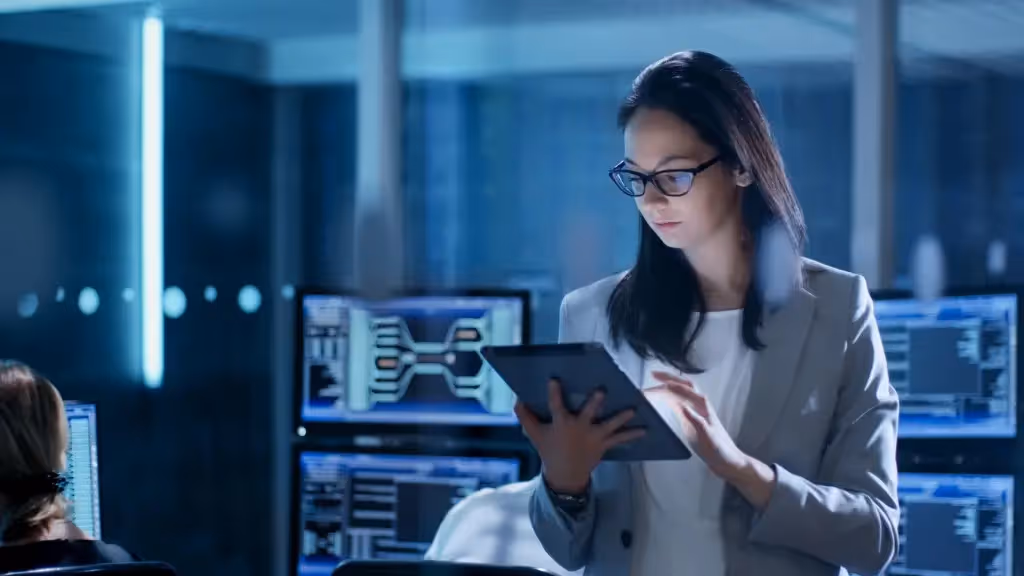 Woman in glasses using a digital tablet in a dimly lit room with multiple computer screens displaying data and diagrams.