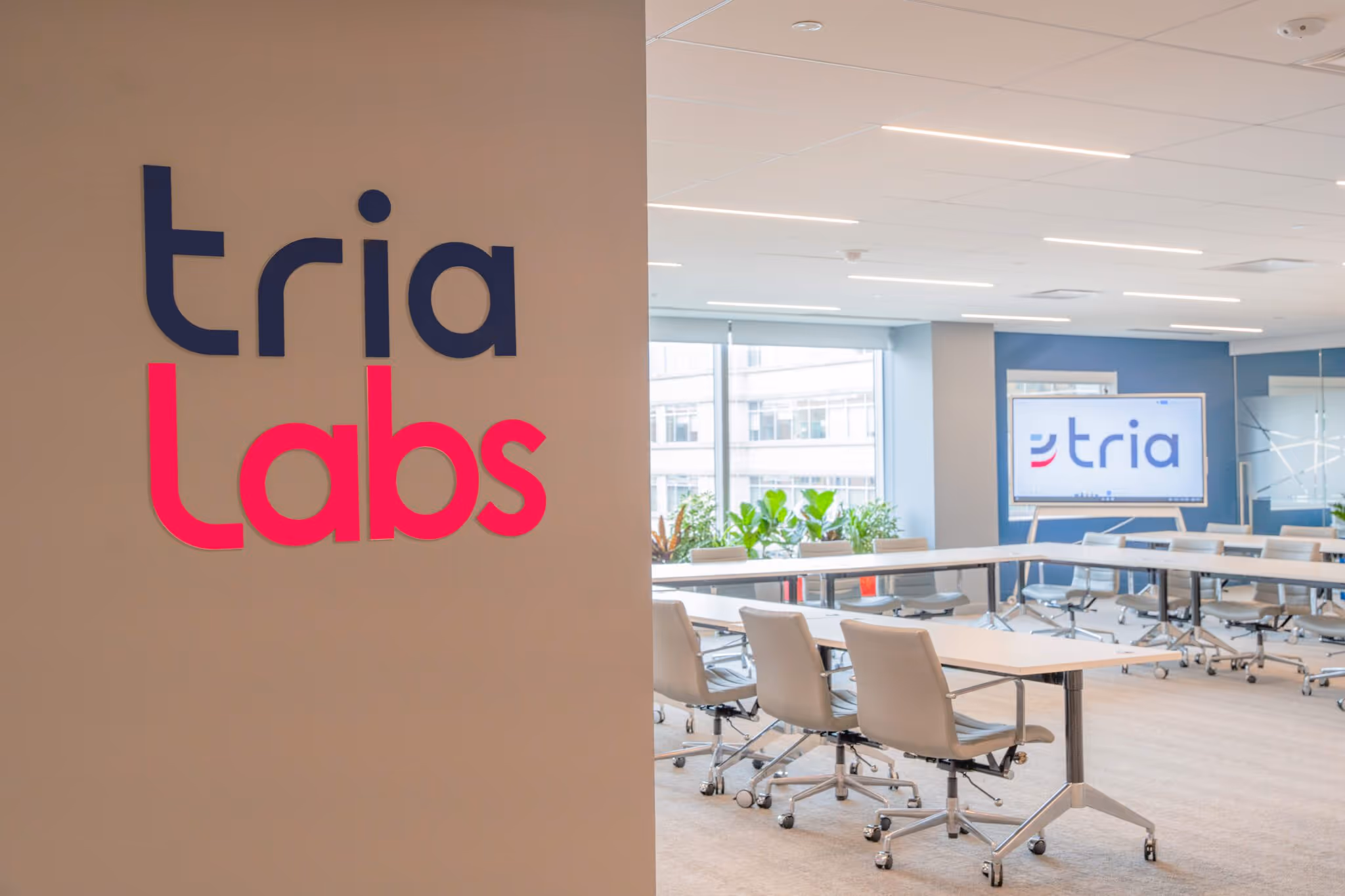 Modern conference room with beige chairs and U-shaped tables, featuring Tria Labs logo on a wall and screen.