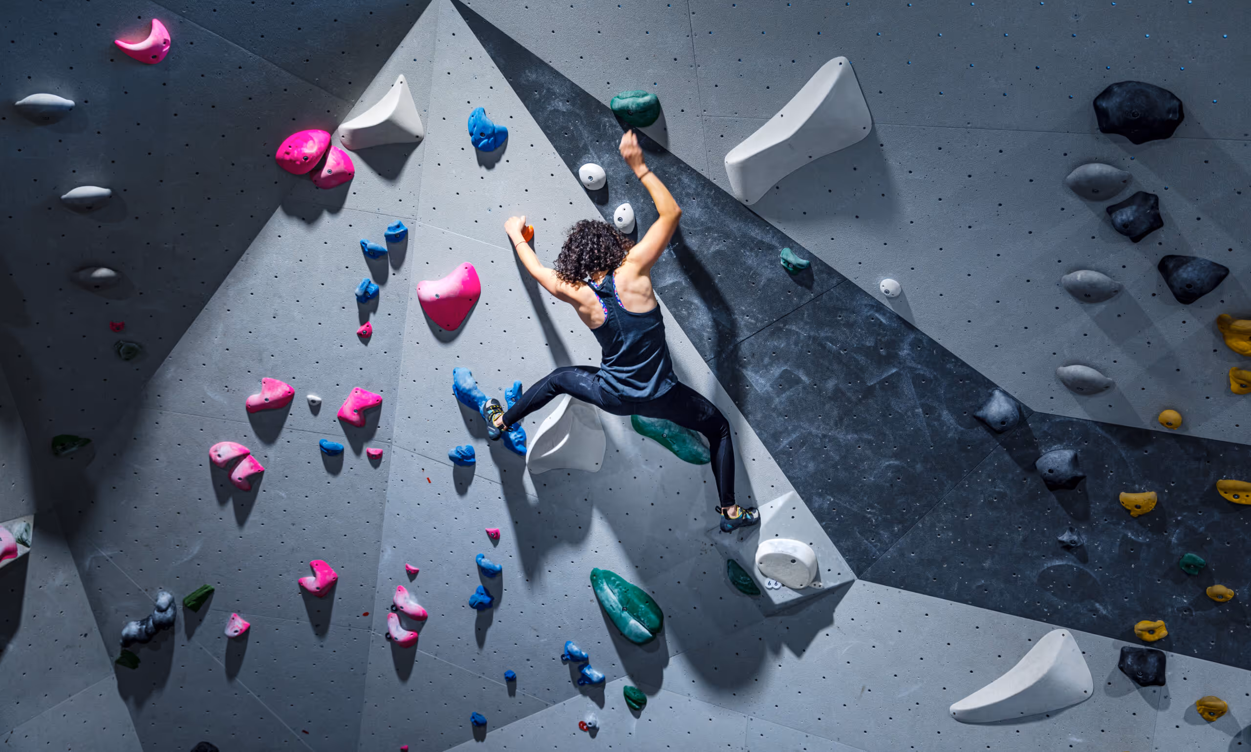 Woman climbing an indoor rock climbing wall with colorful climbing holds in a geometric pattern.