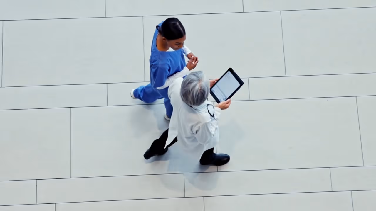 Overhead view of two healthcare professionals walking on a tiled floor, one holding a tablet, with text about Tria Federal's role in federal health systems.