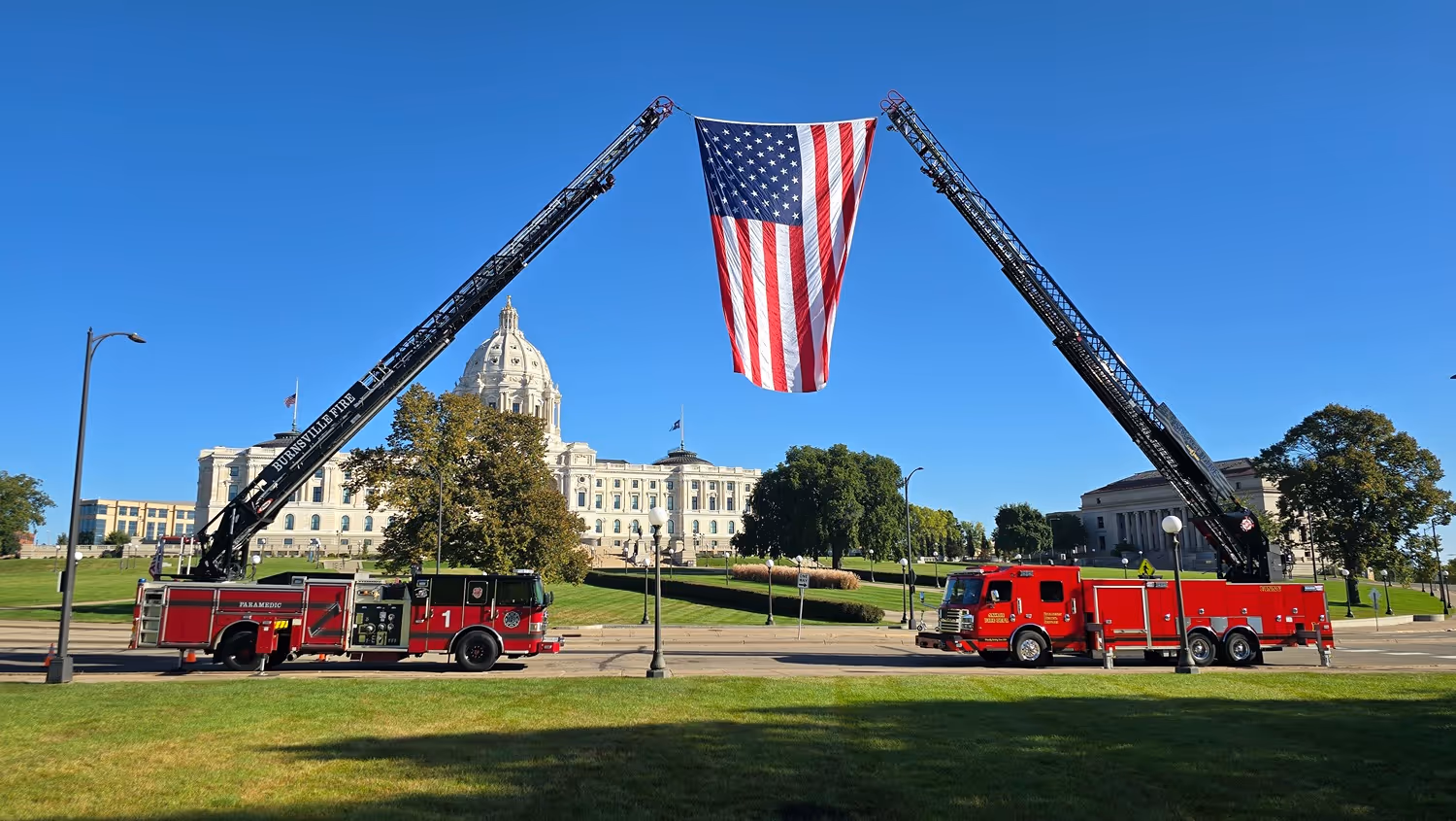 Two fire trucks with extended ladders holding a large American flag in front of a domed government building on a clear day.
