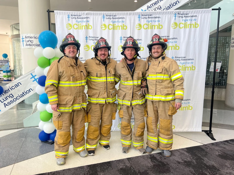 Firefighter wearing full gear and helmet cutting a wooden beam with a power saw, sending sparks flying.