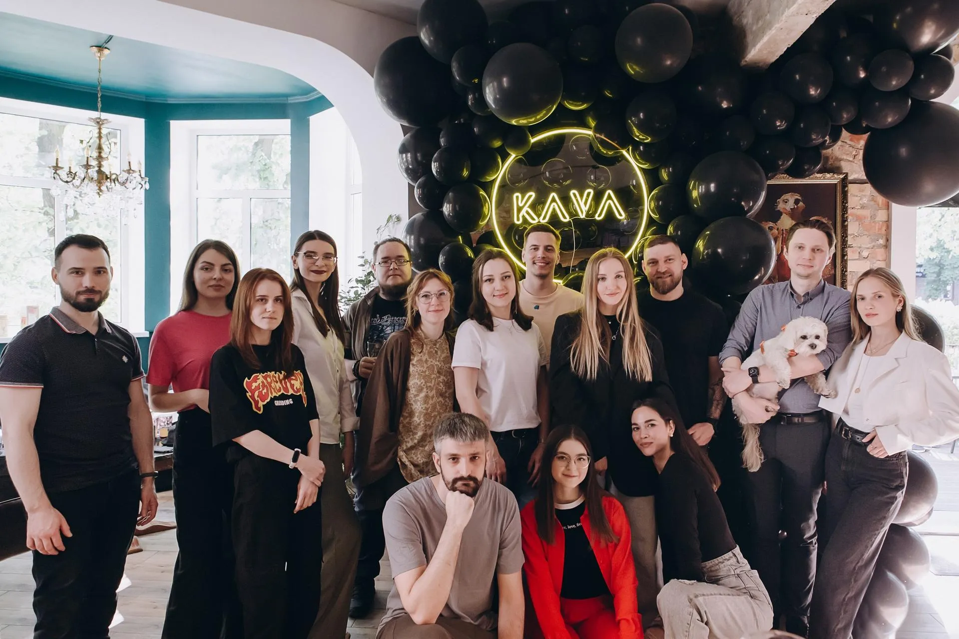 Group photo of about 20 diverse young adults, mostly women in casual trendy outfits like black pants, red tops, and dresses, posing cheerfully inside a stylish cafe or lounge with arched turquoise walls, chandelier, and massive black balloon wall backdrop featuring glowing yellow "KAVA"  neon sign; one man holds a small white dog.

