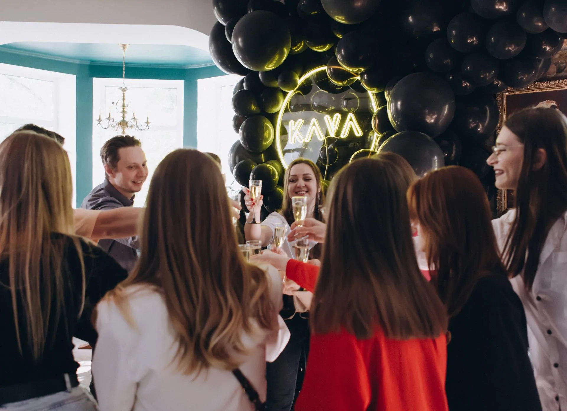Close-up celebratory group photo of eight young adults, primarily women with long hair in white shirts and black pants, joyfully toasting with champagne flutes amid laughter; set against black balloon wall with glowing yellow "KAVA" neon sign in elegant interior with chandelier and arched turquoise walls.
