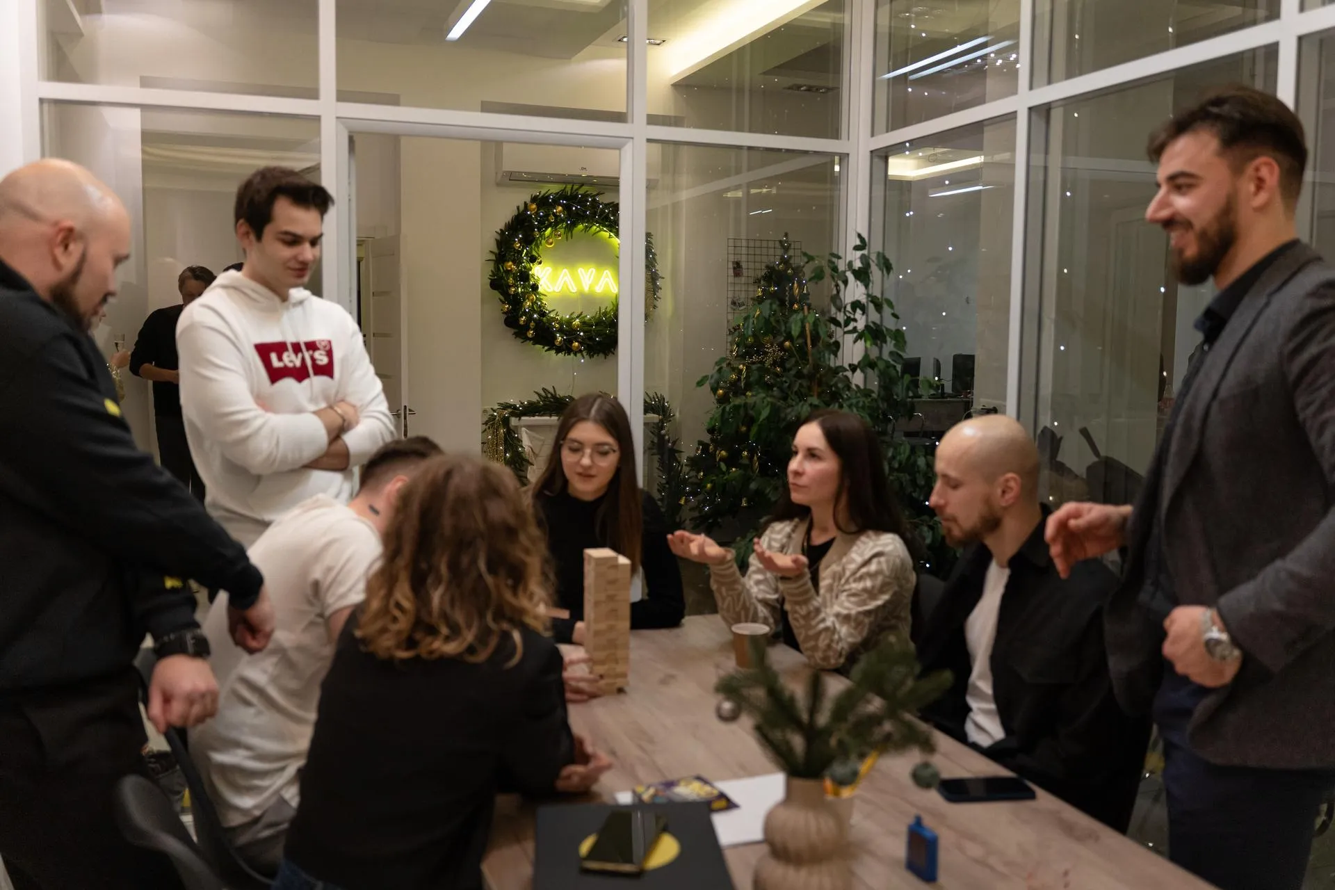 Casual team-building photo of ten young professionals, mix of men and women in hoodies, suits, black clothes, gathered around a wooden table playing Jenga game with holiday decorations; yellow "KAVA" (coffee) neon wreath on glass wall behind small Christmas tree and plants in modern bright office lounge.
