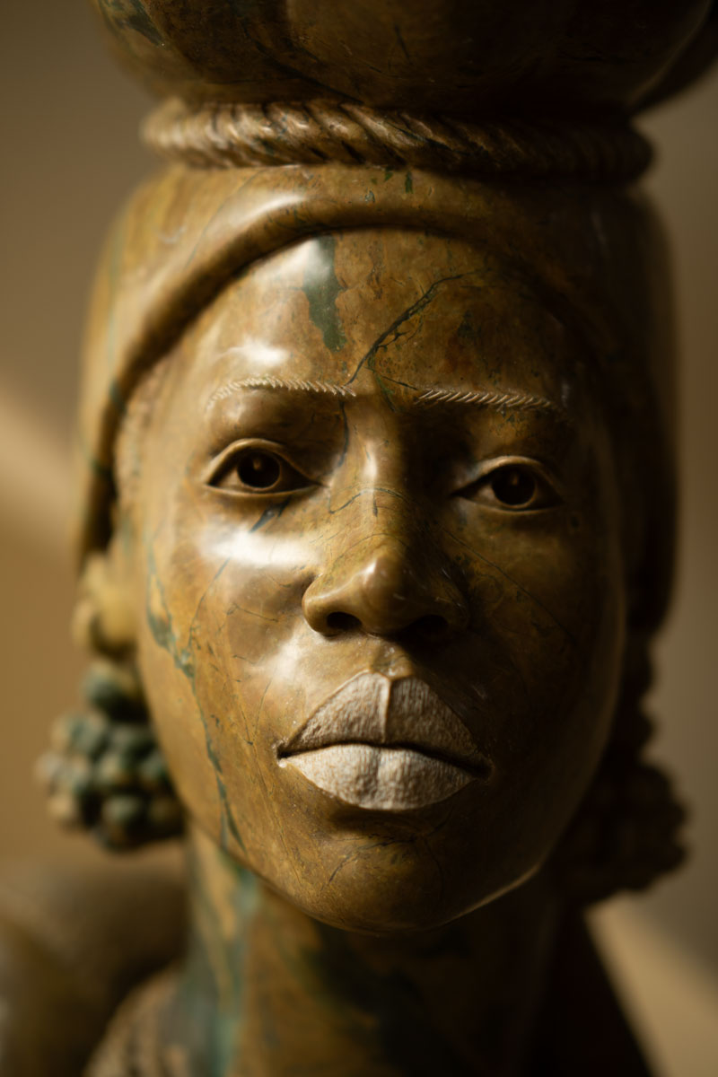Close-up of a detailed stone sculpture of a person's face with carved lips, eyes, and braided hair accessories.
