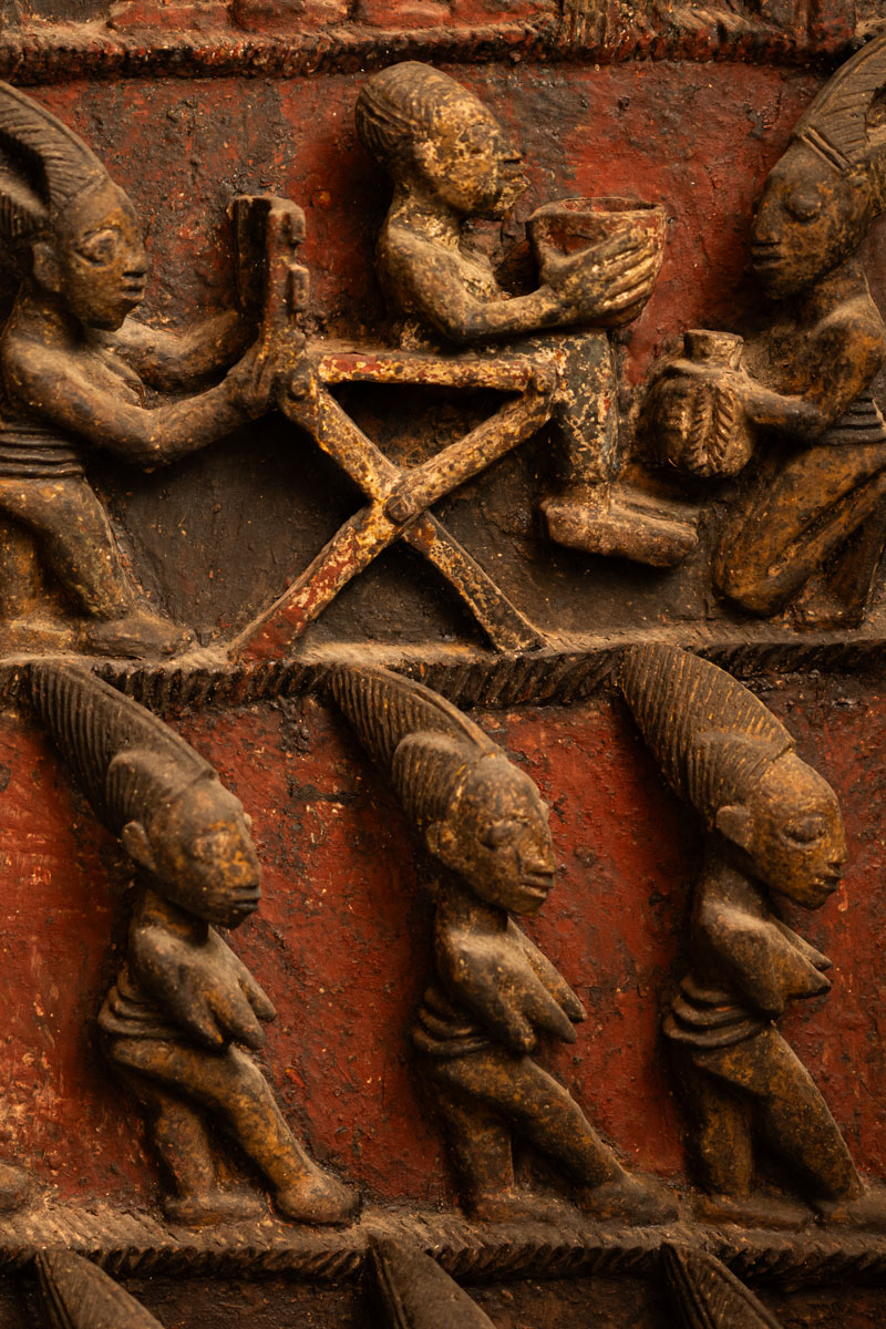 Close-up of a carved wooden relief depicting six human figures with elaborate hairstyles, three standing below and three engaged in various activities above, against a textured reddish background.
