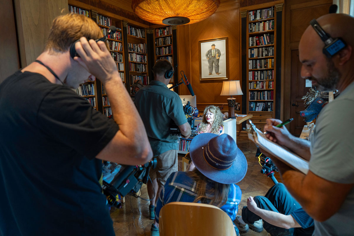 Film crew recording a woman with curly blonde hair seated in a library filled with bookshelves while a crew member takes notes.