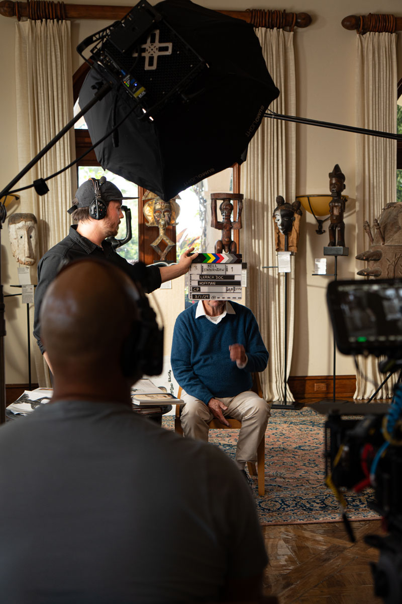 Film crew preparing to shoot an interview in a room decorated with wooden masks and sculptures, with a man seated and a film slate held in front of his face.