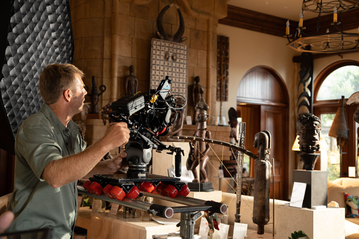 Man operating a professional video camera on a wheeled dolly in a room filled with tribal wooden sculptures and art pieces.