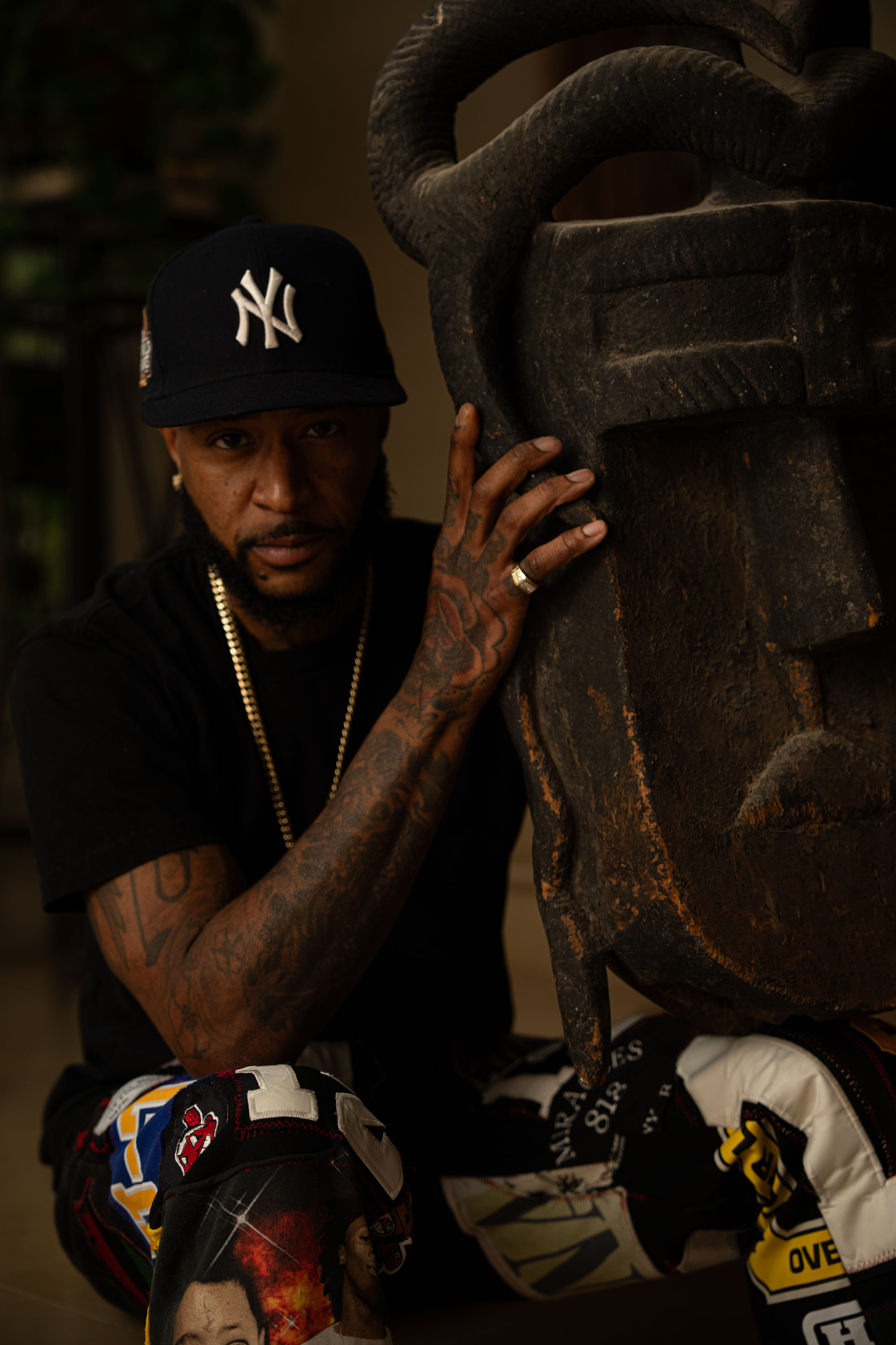 Man wearing a black New York Yankees cap and colorful pants sitting while touching a large pre-modern Toma mask with a horned face.