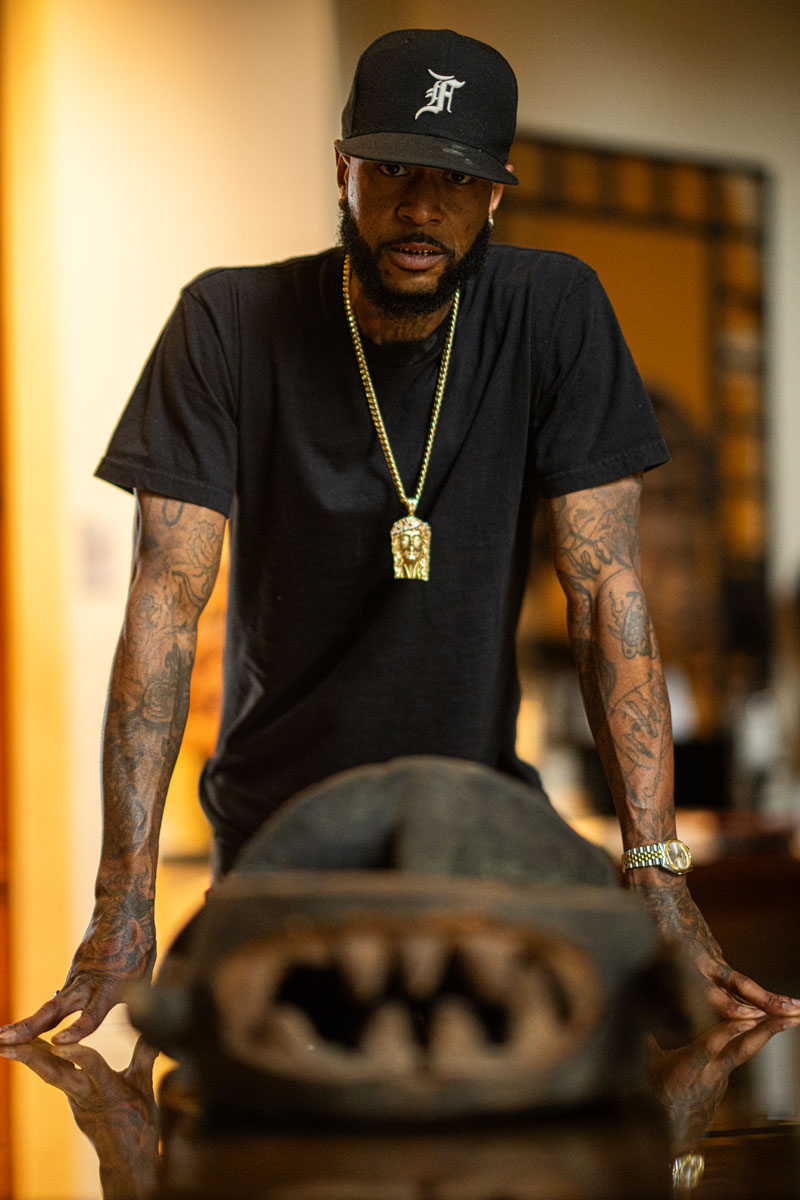 Man in a black t-shirt and cap with tattoos on both arms leaning on a reflective surface with a blurred sculpture in the foreground.