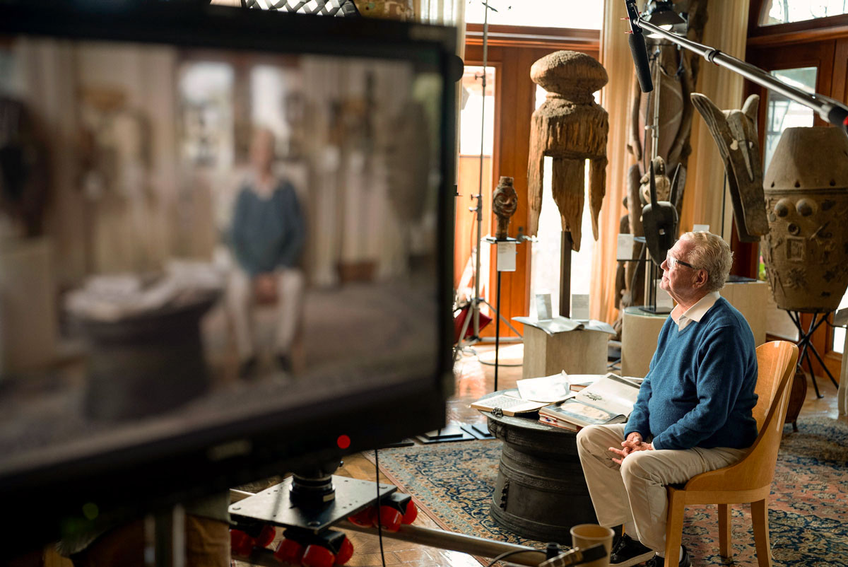 Elderly man wearing glasses and a blue sweater seated on a wooden chair in a room filled with tribal sculptures and artifacts, with a camera monitor showing his blurred image in the foreground.