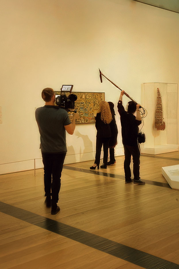 Two museum visitors observing a painting while a film crew records and holds a boom microphone.