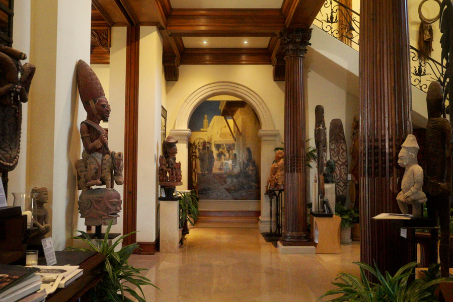 Interior view of an immense private art collection with wooden columns, African tribal sculptures on pedestals, and a mural at the far end.