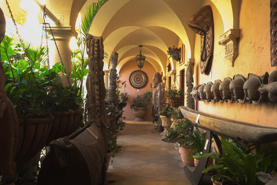 Sunlit corridor with arched ceiling displaying tribal masks, wooden sculptures, and green potted plants.
