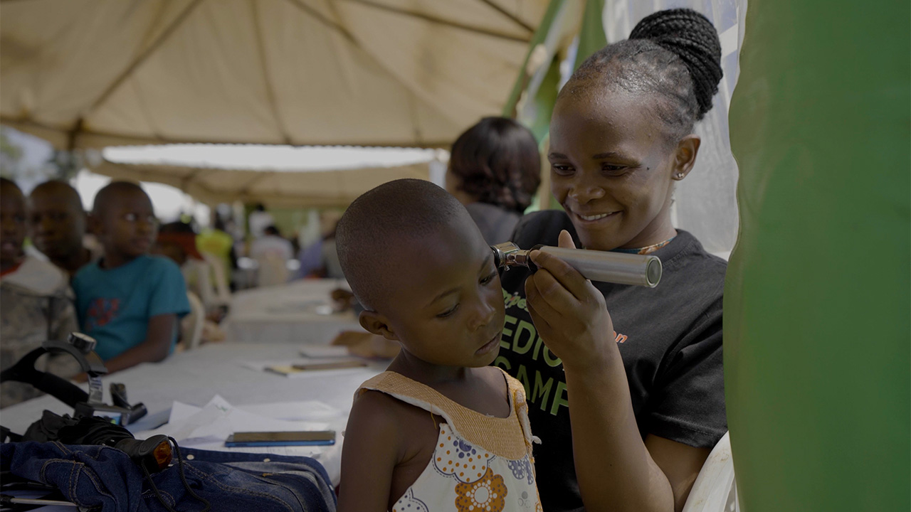 Healthcare worker smiling while examining a young girl's ear with an otoscope under a tent with other children seated in the background.