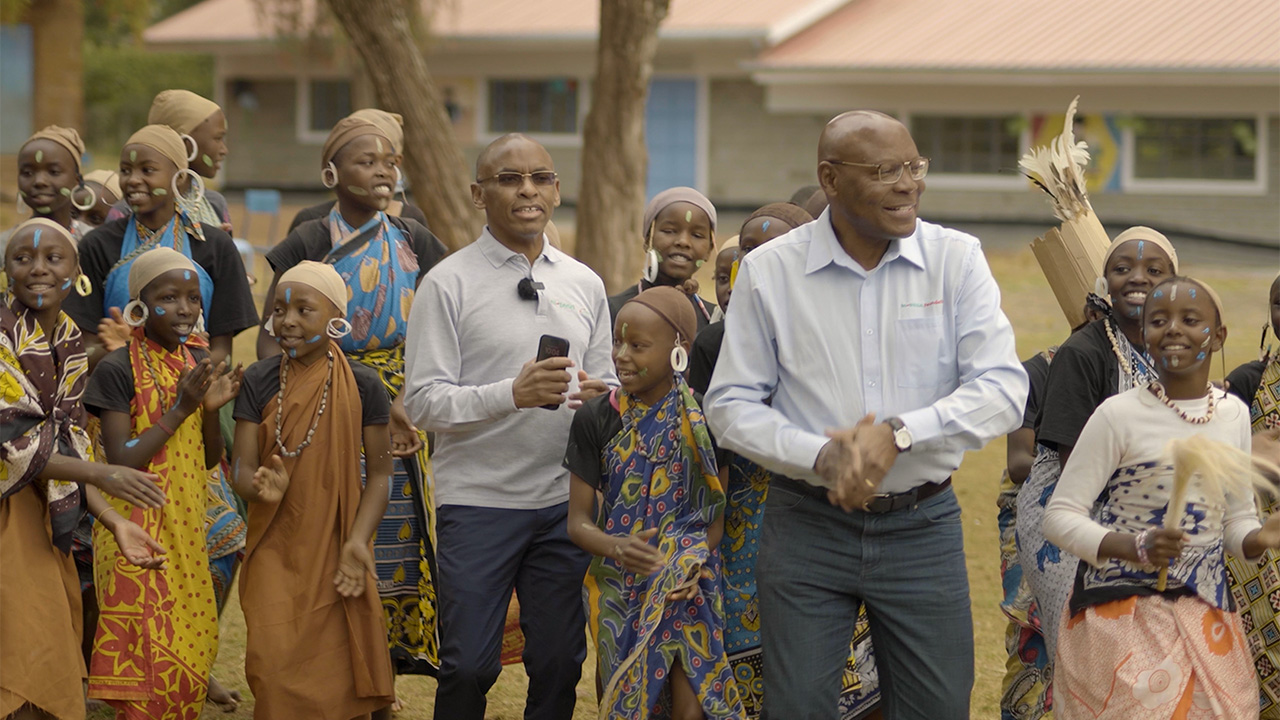 Group of people, including children in colorful traditional attire with face paint and large earrings, dancing outdoors alongside two men in glasses and collared shirts.