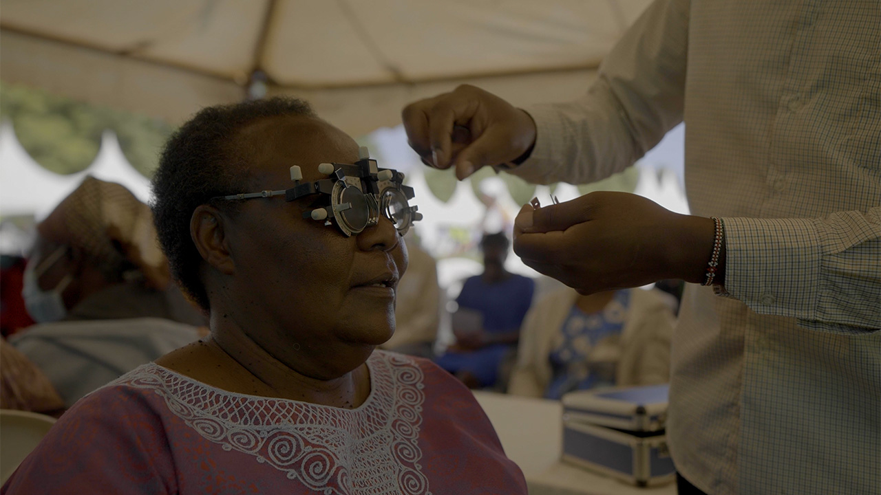 Elderly woman wearing optical testing frames while a person adjusts them in a bright indoor setting.