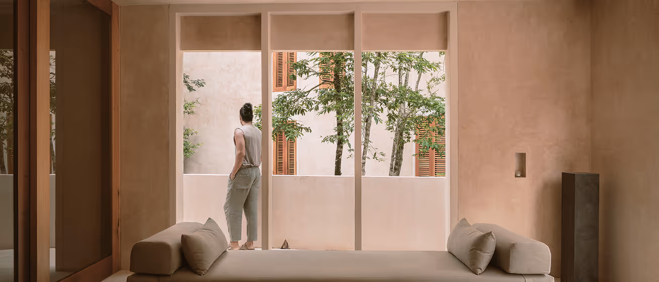 Person standing on a balcony looking at trees through large windows in a minimalist beige room with a cushioned bench.