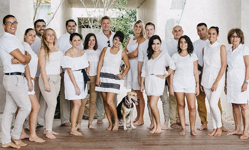 Group of barefoot men and women in white outfits standing on a wooden floor under a concrete structure with a dog in the center.