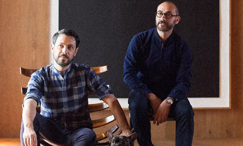 Two men sitting indoors on wooden chairs with a black dog between them.