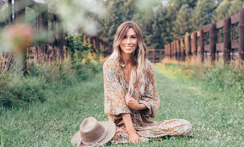 Smiling woman with long blond hair sitting cross-legged on grass next to a beige hat, with wooden fences and trees in the background.